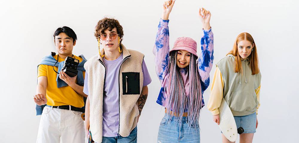 Vier Personen stehen nebeneinander und schauen in die Kamera. Eine Person wirft freudig die Arme nach oben. Zwei weitere Personen tragen ein Skateboard.