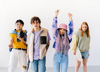 Vier Personen stehen nebeneinander und schauen in die Kamera. Eine Person wirft freudig die Arme nach oben. Zwei weitere Personen tragen ein Skateboard.