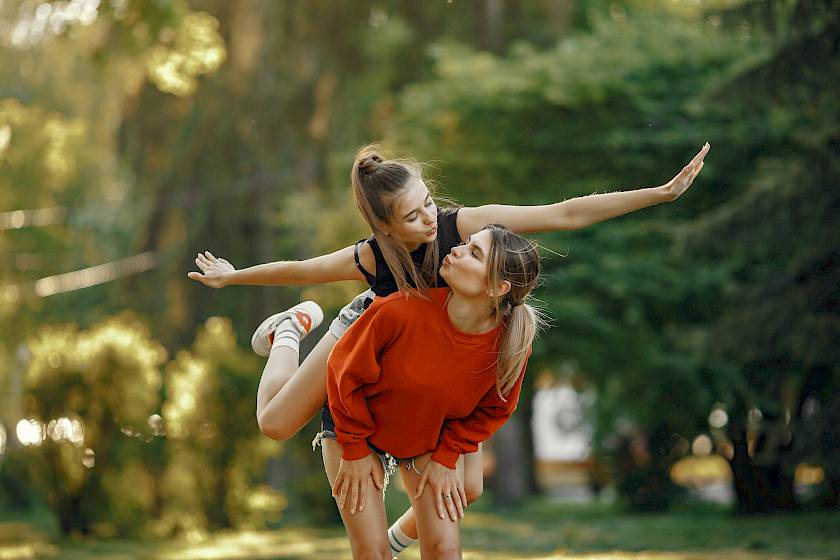 Eine Person sitzt lachend auf dem Rücken einer anderen und streckt die Arme aus wie ein Flugzeug.